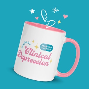 May include: White ceramic mug with a pink interior and handle. The mug features the words "Clinical Depression" in pink script, along with the phrases "Maybe it's" and "Maybe she's born with it." Decorative stars, hearts, and a broken heart graphic are also present.