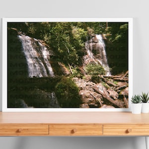 Könnte beinhalten: Ein Foto eines Wasserfalls, der eine felsige Klippe hinunterstürzt, umgeben von üppigem, grünem Laub. Das Wasser ist klar und glitzernd, und das Sonnenlicht erzeugt einen wunderschönen Schein auf der Szene.