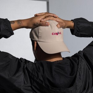 May include: A tan baseball cap with the word "Cuddle" embroidered in pink. The cap is being held by a person wearing a black jacket. The cap has a curved brim and a classic six-panel design.