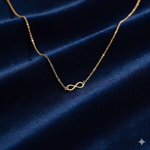 Puede incluir: Un delicado collar dorado con un colgante con el símbolo del infinito. La fina cadena y el colgante destacan sobre un fondo azul oscuro texturizado. Un collar sencillo y elegante, ideal para cualquier ocasión.