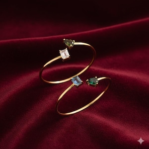 May include: Two gold-toned open cuff bracelets. Each bracelet features a gemstone: one with a white square, one with a blue rectangle, and two with green teardrop-shaped stones. The bracelets are displayed on a deep red velvet surface.