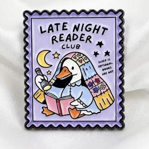 May include: A rectangular enamel pin shaped like a postage stamp with a light purple background. It features a white duck wearing a blanket, holding a flashlight and an open book. Text reads: LATE NIGHT READER CLUB and SLEEP IS OPTIONAL, BOOKS ARE NOT.