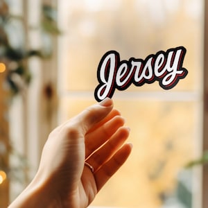 May include: A sticker with the word "Jersey" in a cursive font. The word is white with a red outline and a black border. The sticker is held in a hand with a gold ring.