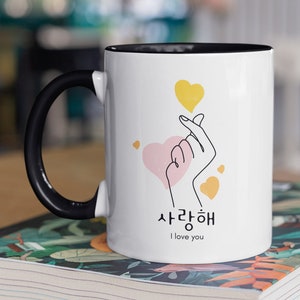 May include: White ceramic mug with a black handle. The mug has a pink heart with a hand making a heart sign. The mug also has yellow hearts and the text "사랑해" which translates to "I love you" in English.