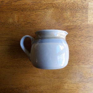 May include: A small, white ceramic creamer with a light brown rim and a light blue band around the middle. The creamer has a rounded body and a handle.
