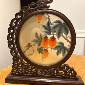 May include: A dark brown, intricately carved wooden display stand. It features a circular painting of orange fruit and green leaves. The stand has a dragon head at the top and scrollwork. The painting is set within a dark brown circular frame.