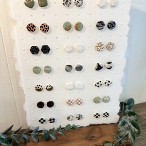 Op de afbeelding: Een display met 20 paar keramische oorbelletjes met verschillende patronen en kleuren, waaronder zwart-wit, groen-wit, goud-wit en bruin-wit. De oorbelletjes worden getoond op een wit bord met een houten basis.