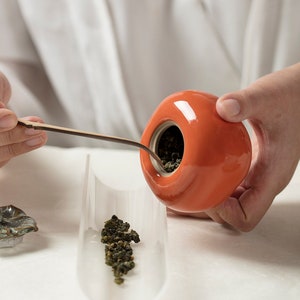 May include: A person is pouring loose leaf tea from an orange ceramic tea pot into a clear glass tea infuser. The tea leaves are green and brown.