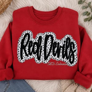 May include: A red crewneck sweatshirt featuring the text Red Devils in black script lettering. The text is layered over a white patch with black polka dots. The words SBD Embroidery are printed in small white script below the main design.