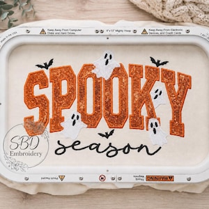May include: An 8x13 inch embroidery hoop containing off-white fabric with the word SPOOKY in large orange glittery block letters and the word season in black cursive script. White ghost and black bat appliques surround the text.