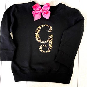 May include: Black sweatshirt with a large leopard print letter "G" on the front. A pink bow is on top of the sweatshirt.