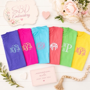 May include: Six short-sleeved t-shirts in purple, hot pink, light blue, coral, lime green, and yellow, each featuring a unique embroidered monogram on the chest. A sign reads SBD Embroidery and another card reads monogram MADE JUST FOR YOU.