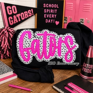 May include: A black sweatshirt featuring a large, central embroidered applique with the word "Gators" in bright pink glittery script. The text is layered over a white background with black polka dots and a scalloped edge, creating a textured look.