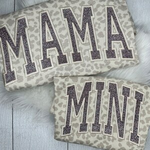 May include: Two matching sweatshirts, one large and one small, with a light beige leopard print. The word "MAMA" is embroidered on the larger sweatshirt, and "MINI" is embroidered on the smaller one, both in a glittery, dark brown font.