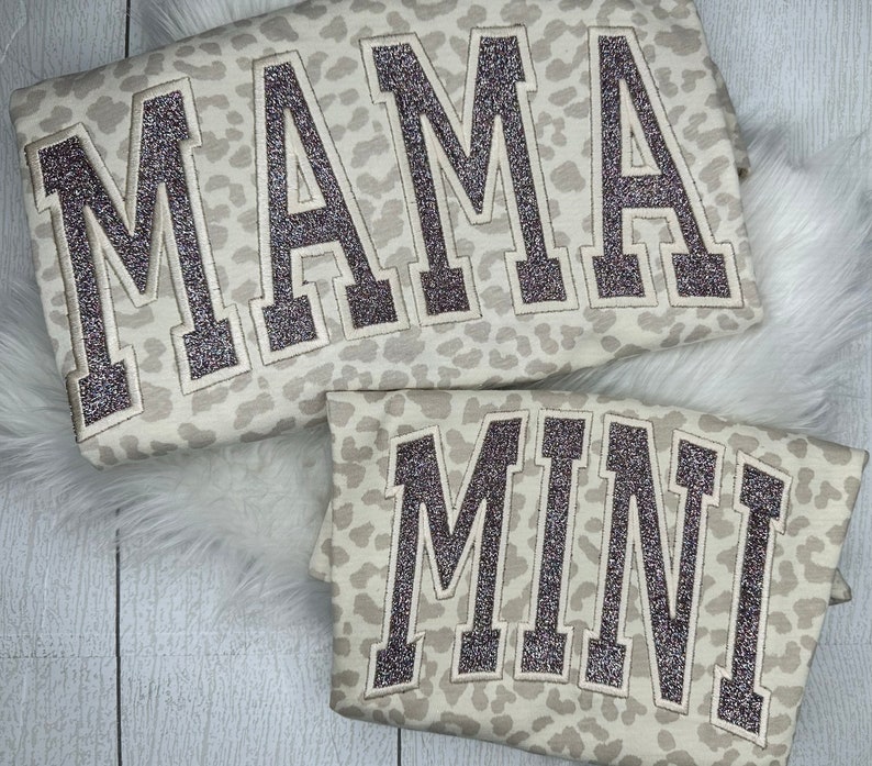 May include: Two matching sweatshirts, one large and one small, with a light beige leopard print. The word "MAMA" is embroidered on the larger sweatshirt, and "MINI" is embroidered on the smaller one, both in a glittery, dark brown font.