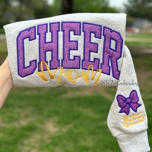 May include: Gray sweatshirt with purple glitter "CHEER MOM" embroidery and a purple glitter bow on the sleeve. The text "SBD Embroidery" is also embroidered on the sleeve.