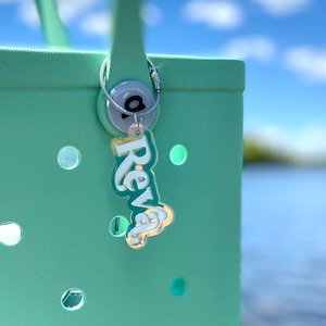 May include: A light green tote bag with a silver key chain attached. The key chain has a silver charm with the word "Reva" written in white letters.