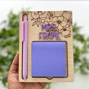 May include: A wooden notepad holder with a floral design and the text "MRS FRAME". It includes a purple pen and a square of purple sticky notes. The holder is held in a hand.