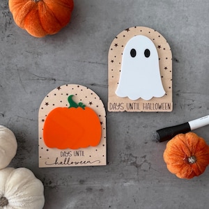 May include: Two Halloween countdown signs with a pumpkin and ghost design. The signs are made of wood and acrylic, with the words "Days Until Halloween" printed on them. Decorative pumpkins and a marker are also in the image.