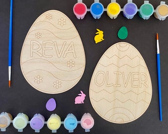 May include: Two wooden Easter egg shapes with the names "Reva" and "Oliver" written on them. The eggs are surrounded by paint pots, paintbrushes, and small bunny and egg cutouts.