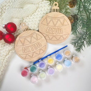 May include: Two unfinished wooden Christmas ornament shapes with the names "Reva" and "Nitya" written on them. The ornaments are surrounded by paint pots and a paintbrush.