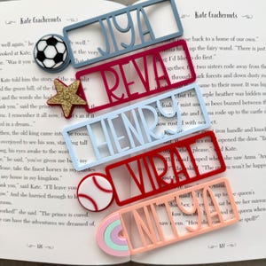 Può includere: Una collezione di segnalibri colorati a forma di nome e spille decorative, tra cui un pallone da calcio, una stella, una palla da baseball e un arcobaleno. I segnalibri scandiscono nomi come Jiya, Reva, Henry e Mira.