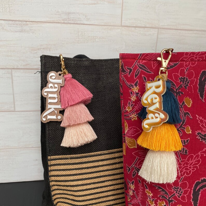 May include: Two personalised keychains with tassels. The keychain on the left says "Janie" in white letters on a brown background. The keychain on the right says "Reva" in white letters on a gold background. Both keychains have three-tiered tassels in different colours.