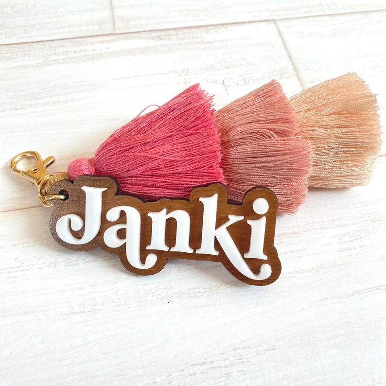 May include: A wooden keyring with the name "Janki" in white lettering. The keyring has a pink and beige tassel attached to a gold metal ring.