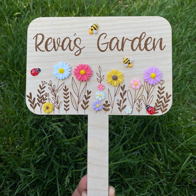 Pu&ograve; includere: Un cartello da giardino in legno con il testo "Reva's Garden" inciso su di esso. Il cartello &egrave; decorato con piccoli fiori e coccinelle di plastica.