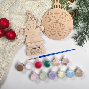 May include: A gingerbread person and a Christmas ornament, both unfinished and ready to be painted. The ornament is round and has a geometric design with the name "Reva" written on it. The gingerbread person is holding a sign with the same name. The set includes 12 paint pots in various colors and a paintbrush.