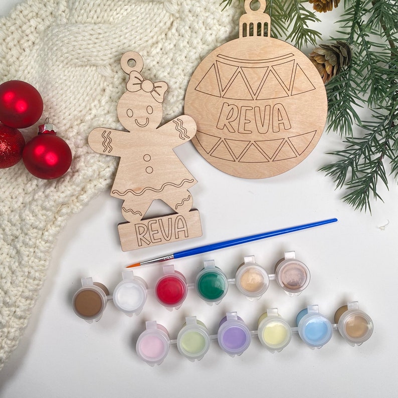 May include: A gingerbread person and a Christmas ornament, both unfinished and ready to be painted. The ornament is round and has a geometric design with the name "Reva" written on it. The gingerbread person is holding a sign with the same name. The set includes 12 paint pots in various colors and a paintbrush.