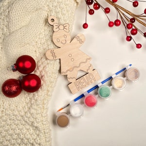 May include: A wooden gingerbread person ornament with the name "Reva" on the base. The ornament is unpainted and comes with six small paint pots and a paintbrush.