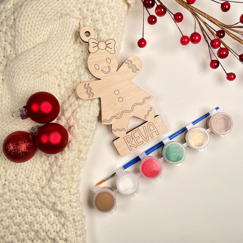 May include: A wooden gingerbread person ornament with the name "Reva" on the base. The ornament is unpainted and comes with six small paint pots and a paintbrush.