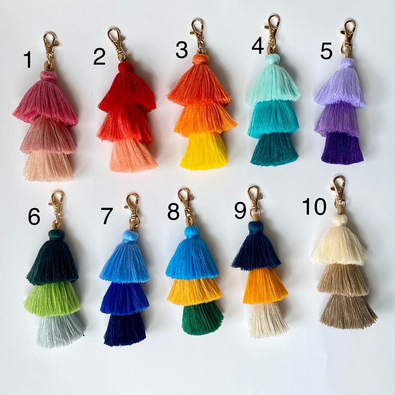 May include: Ten colourful tassel keychains with three layers of fringe. The tassels are in various colours, including pink, red, yellow, blue, green, and purple.