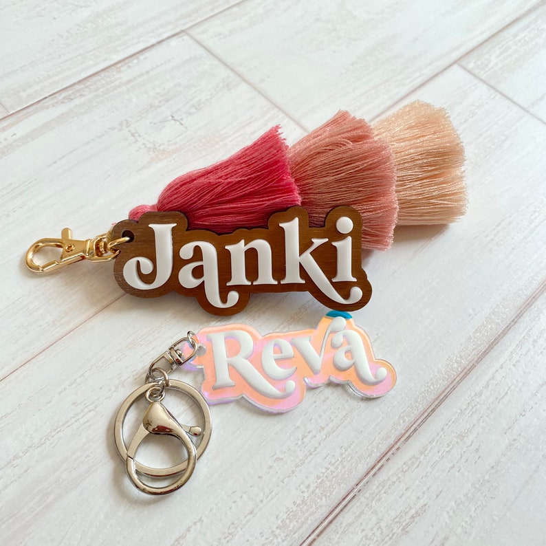 May include: Two personalised keychains with the names "Janki" and "Reva". The "Janki" keychain is made of wood with a pink and peach tassel. The "Reva" keychain is made of clear acrylic with a pink and peach iridescent finish.