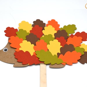 May include: A brown paper hedgehog with a googly eye and a red pom-pom nose. The hedgehog is covered in colorful paper leaves in shades of red, orange, yellow, green, and brown.