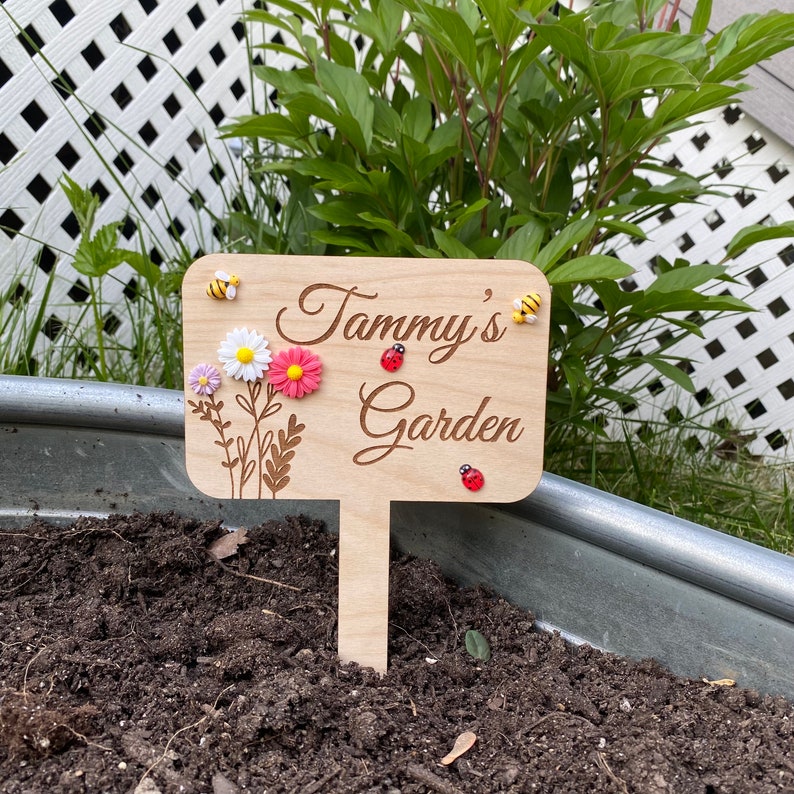 Pu&ograve; includere: Un cartello da giardino in legno con il testo "Tammy's Garden" inciso su di esso. Il cartello &egrave; decorato con piccoli fiori colorati e coccinelle.