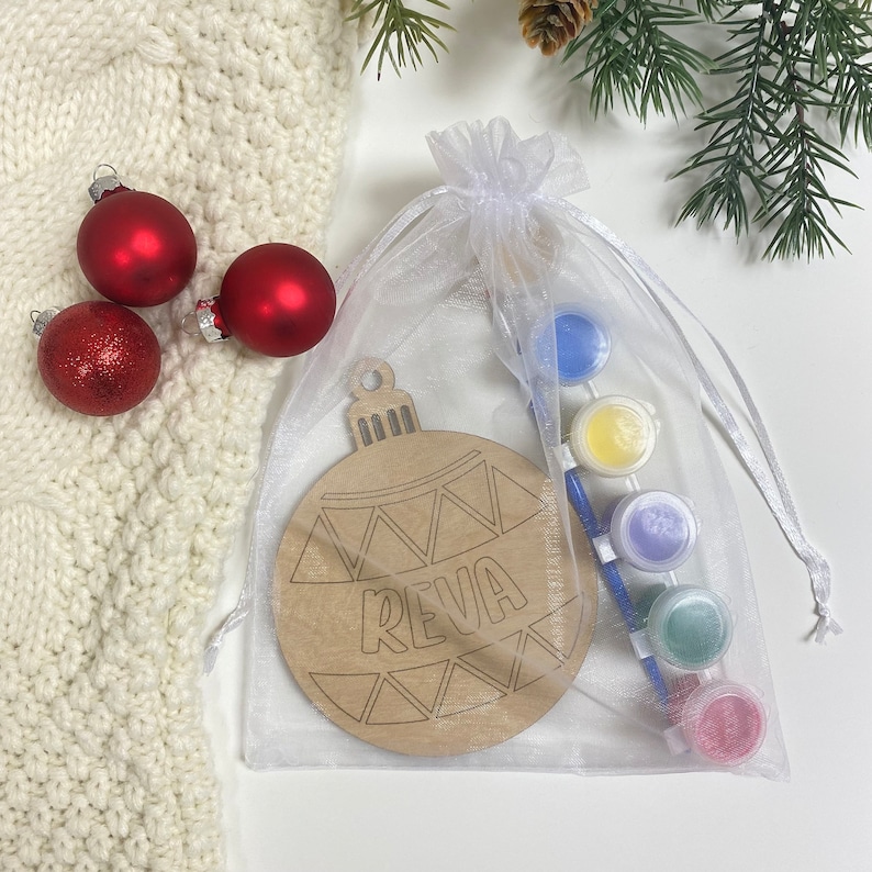 May include: A white organza bag filled with paint and a paintbrush, a wooden ornament shaped like a Christmas tree with the name "Reva" written on it, and three red Christmas ornaments.