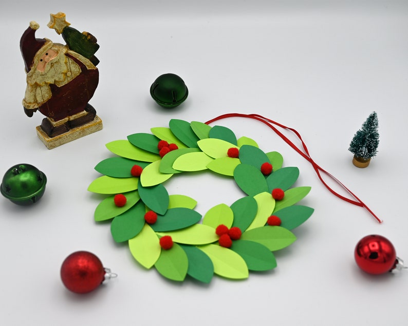 May include: A green paper Christmas wreath with red pom-poms, a red ribbon, and a small Christmas tree figurine.