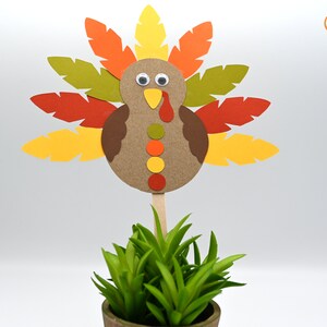 May include: A paper craft turkey with brown, orange, yellow, and green feathers. The turkey has googly eyes and a yellow beak. The turkey is on a stick and is in a pot with green grass.