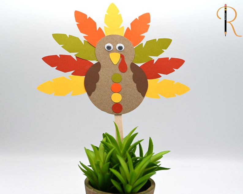 May include: A paper craft turkey with brown, orange, yellow, and green feathers. The turkey has googly eyes and a yellow beak. The turkey is on a stick and is in a pot with green grass.