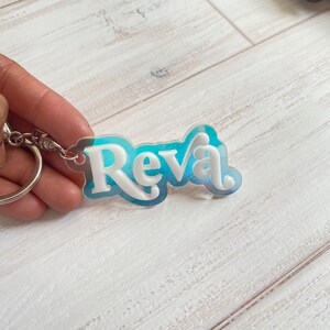 May include: A white acrylic keychain with the name "Reva" in a blue and white gradient. The keychain is attached to a silver metal ring.