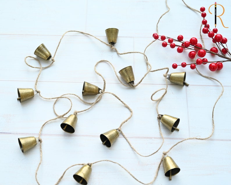 May include: A string of small gold bells hanging from a twine string. The bells are evenly spaced and are all the same size. The string is hanging from a white surface.