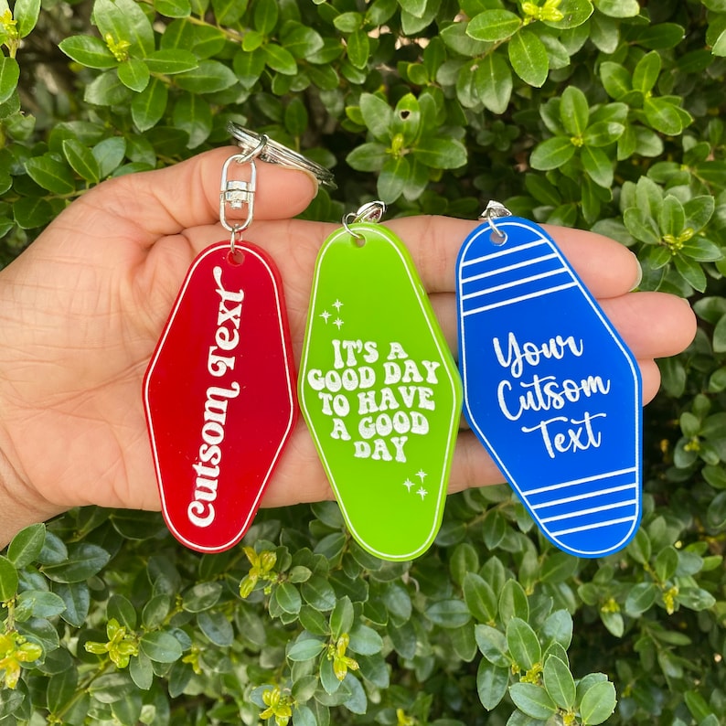 Op de afbeelding: Drie motel sleutelhanger sleutelhangers met verschillende kleuren en tekst. De rode sleutelhanger zegt "custom text". De groene sleutelhanger zegt "It's a good day to have a good day". De blauwe sleutelhanger zegt "Your custom text".