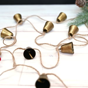 May include: A string of small gold bells connected by twine. The bells are hanging on a white surface.