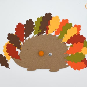 May include: A brown paper hedgehog with a googly eye and an orange pom-pom nose. The hedgehog is surrounded by fall-colored paper leaves in shades of red, orange, yellow, and green.