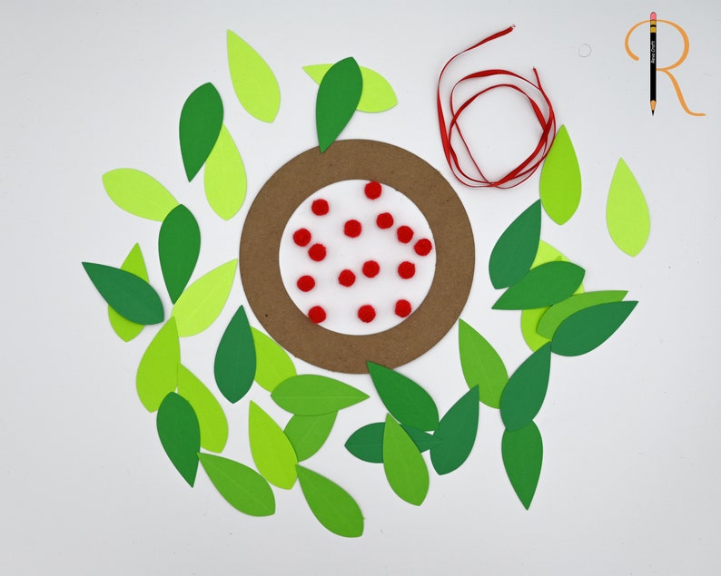 May include: A brown paper circle with red pom-poms on a white background surrounded by green and light green paper leaves. A red ribbon is also included.