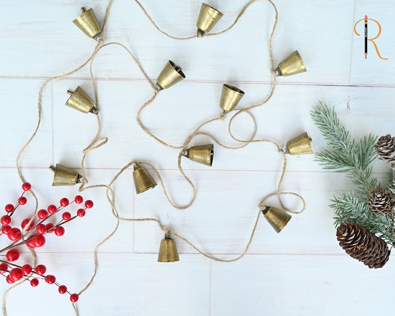 May include: A string of gold bells hanging on a white background. The bells are connected by a natural twine string.