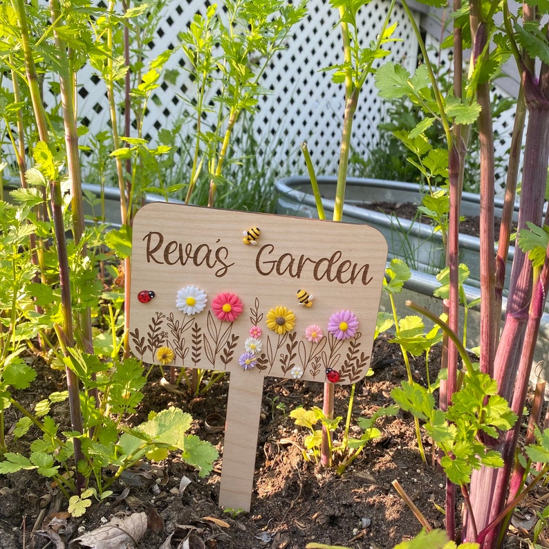 Pu&ograve; includere: Un cartello da giardino in legno con il testo "Reva's Garden" e fiori e api decorative. Il cartello &egrave; posizionato in un giardino con piante verdi.