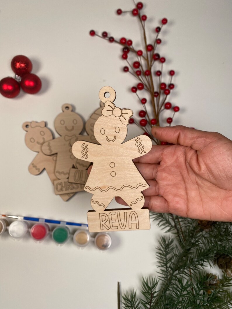 May include: Unfinished wooden gingerbread person ornament with a bow and the name "Reva" written on the base. The ornament is surrounded by paint, paintbrushes, and Christmas greenery.
